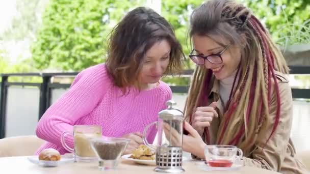 Menina moderna com longos dreadlocks está mostrando algo no smartphone para sua bela amiga em suéter rosa durante uma pausa para almoço juntos em uma cafeteria ao ar livre
 - Filmagem, Vídeo