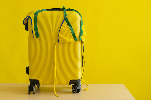 Een openhartige mini-bikini van de kleur van de Braziliaanse vlag hangt op een koffer op een gele achtergrond. Het concept van een zomervakantie. Een vrouw gaat op reis naar de zee.. - Foto, afbeelding