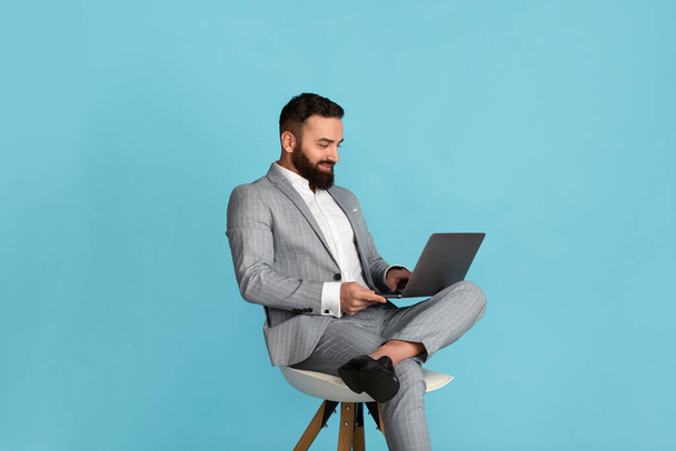 Homme d'affaires attrayant utilisant un ordinateur portable tout en étant assis sur une chaise sur fond bleu - Photo, image