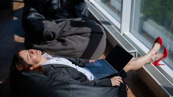 Business lady sleeps sitting in a modern office comfortable bob chair bag. A woman in a suit fell asleep at work on a laptop. Caring for the convenience and comfort of employees. - Foto, imagen