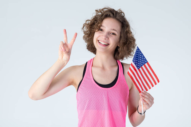 Happy young girl holding USA flag on a gray background. Woman showing sign V. - Fotó, kép