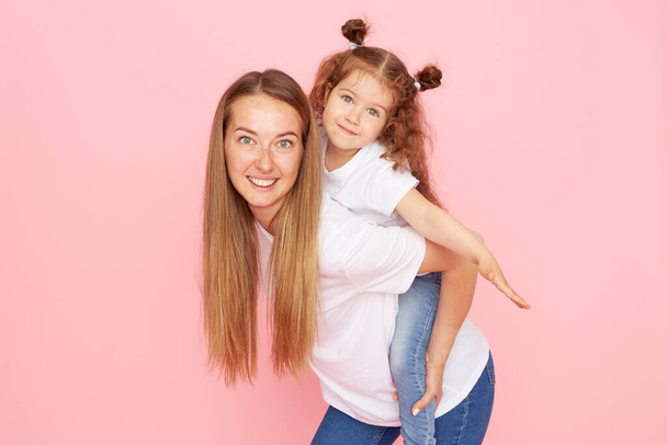 Mom and daughter in white t-shirts and jeans play and hug on a pink background. Caring for loved ones. Happy motherhood - Photo, Image