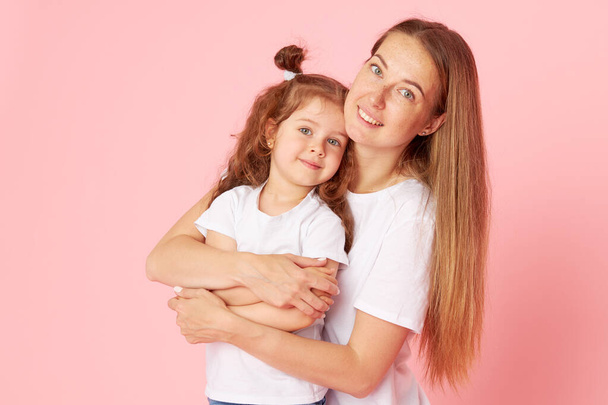 Mom and daughter in white t-shirts and jeans play and hug on a pink background. Caring for loved ones. Happy motherhood - 写真・画像