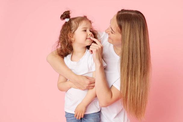 Mom and daughter in white t-shirts and jeans play and hug on a pink background. Caring for loved ones. Happy motherhood - Foto, immagini