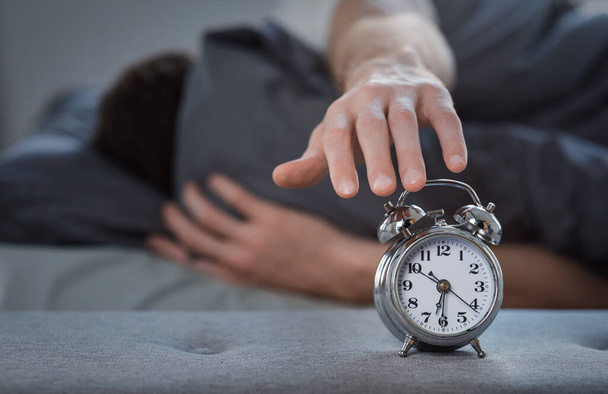 Человек выключает будильник, лежащий в постели дома
 - Фото, изображение