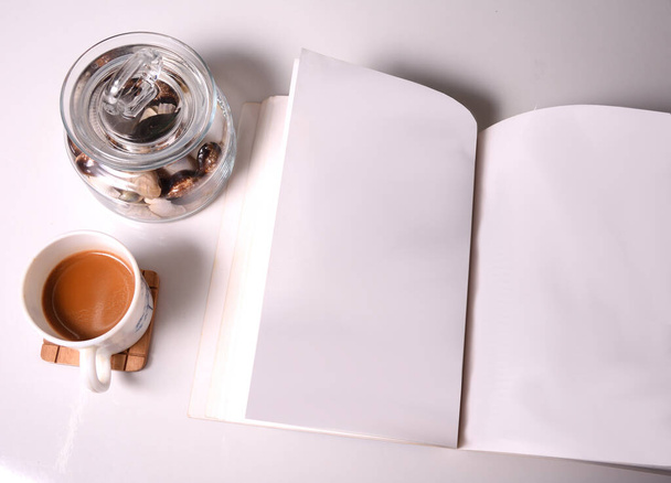 Filiżanka herbaty na białym stole wraz z pustą książką i szklanym pojemnikiem wypełnionym muszlami morskimi  - Zdjęcie, obraz