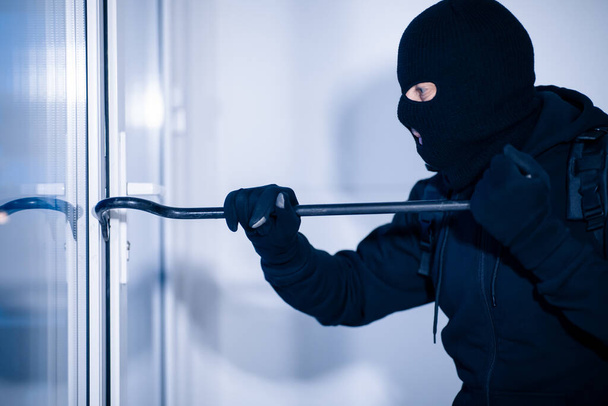 Robber dans la porte de fissuration de cagoule noire avec le pied de biche - Photo, image