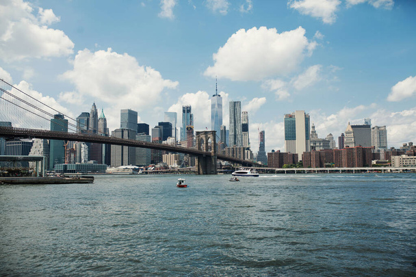 Cityscape Hudson River és Brooklyn Bridge hajók és jachtok ellen kék ég felhők, felhőkarcolók és nagy épületek acél és üveg New Yorkban. Tájnap megalopoliszban - Fotó, kép