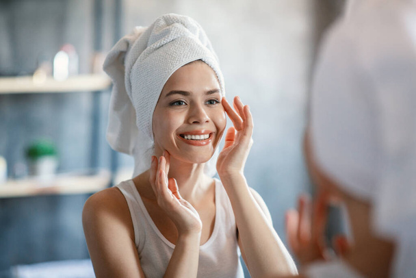Ευτυχισμένη χιλιετή γυναίκα με πετσέτα στο κεφάλι και λαμπερό δέρμα μπροστά στον καθρέφτη στο μπάνιο - Φωτογραφία, εικόνα
