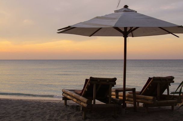 Trópusi esernyő és bambusz hosszú székek a tengerparton, nyugodt tenger naplementekor, Gili Air Island, Lombok, Indonézia - Fotó, kép