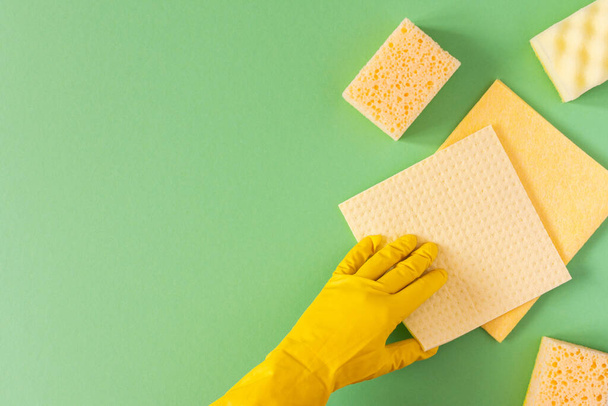 Рука в желтой резиновой перчатке с моющими губками на зеленом фоне
 - Фото, изображение