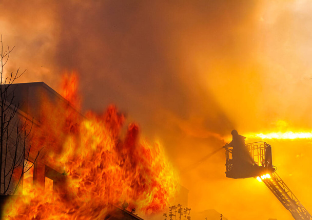 Silhueta de bombeiro em uma escada de elevação de guindaste extinguindo muitas chamas de uma casa em chamas com telhado plano. Imagem dramática com fogo e pôr do sol no céu laranja e amarelo. Imagem com espaço de cópia
. - Foto, Imagem