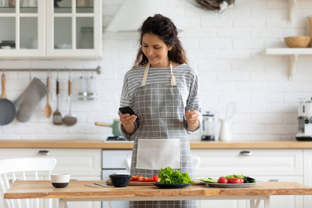 Felice giovane donna attraente cucina alla ricetta su smartphone. - Foto, immagini
