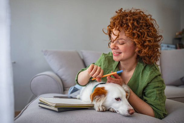 A girl writes in a notebook and plays with a dog. Redhead woman smiling, hugging with dog at home. Shelter a mongrel at home. Life style. College People Study Learning Reading Lecture Notes - Photo, image