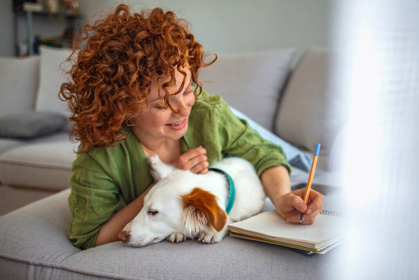 A girl writes in a notebook and plays with a dog. Redhead woman smiling, hugging with dog at home. Shelter a mongrel at home. Life style. College People Study Learning Reading Lecture Notes - Foto, afbeelding