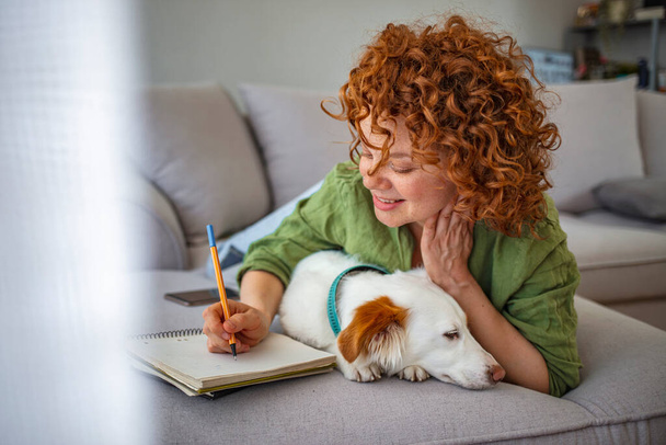Κοκκινομάλλα γυναίκα που δουλεύει στο σπίτι με το σκύλο της. Πορτρέτο μιας όμορφης γυναίκας με το σκύλο της στο σπίτι. Ξαπλωμένη κοπέλα χαλαρώνει στον καναπέ στο σπίτι. Αναψυχή και άνεση έννοια άνθρωποι.  - Φωτογραφία, εικόνα