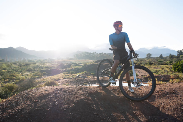 backlit image of a mountain biker and his bike on top of a mound with a mountain range in the distance - Photo, Image