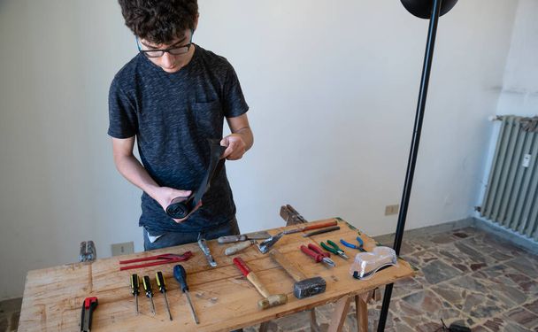Um menino caucasiano tem a intenção de encomendar ferramentas de trabalho em uma mesa: chaves de fenda, martelos, alicates, faca de utilidade e óculos de proteção podem ser vistos
. - Foto, Imagem