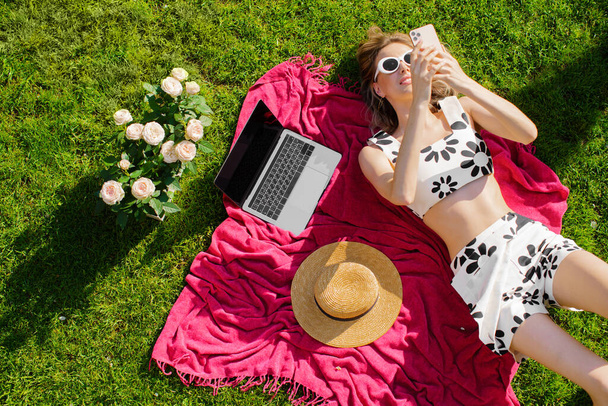 Young pretty woman in sunglasses lying on lawn surfing Internet with laptop, hat and roses, photo for blog or ad - Foto, Imagem