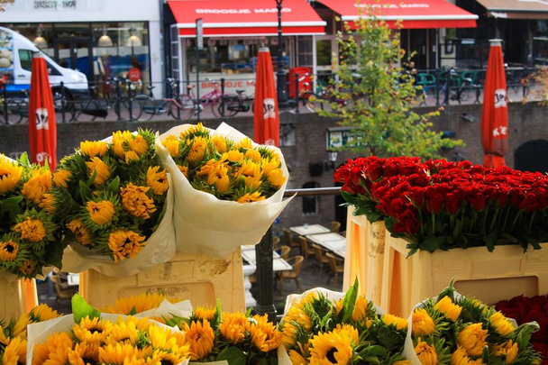 UTRECHT, NETHERLANDS - OCTOBER 20. 2018: View on bouquets of yellow sunflowers and red tulips near water canal on flower market - Фото, изображение