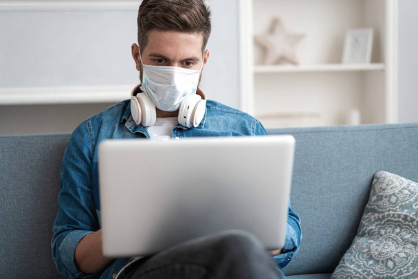 Young caucasian man working from home, wearing protective mask, using laptop. Coronavirus pandemic, covid 19 quarantine - Φωτογραφία, εικόνα