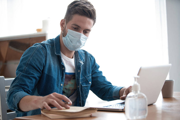 Молодой кавказский человек работает из дома, носит защитную маску, использует ноутбук. Пандемия коронавируса, ковид 19 карантин
 - Фото, изображение
