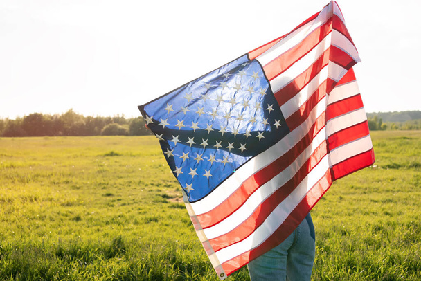Женщина, держащая американский флаг над головой на парковом поле во время заката. Вид сзади
. - Фото, изображение