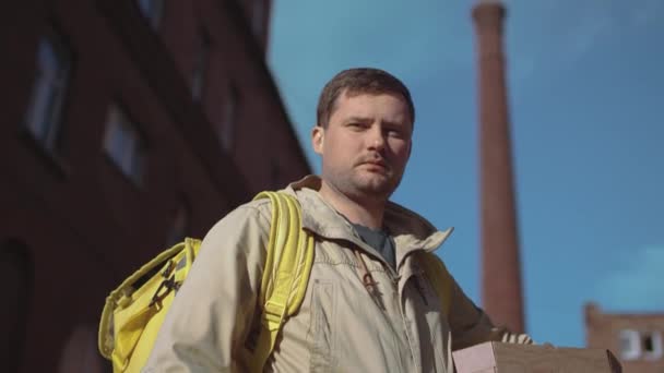 Repartidor caucásico en una chaqueta con una mochila amarilla y cajas de pizza de pie en la calle en un clima soleado y mirando a la cámara
. - Imágenes, Vídeo