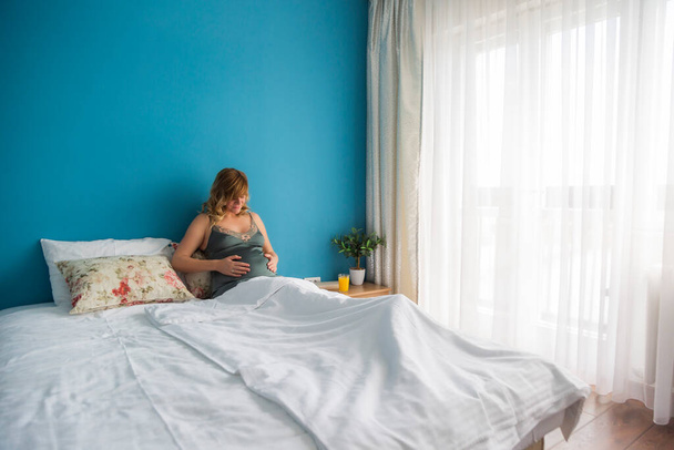 Una mujer embarazada sonriente en una posición semi-sentada en la cama con las manos alrededor del estómago. Está mirando hacia su estómago.. - Foto, Imagen