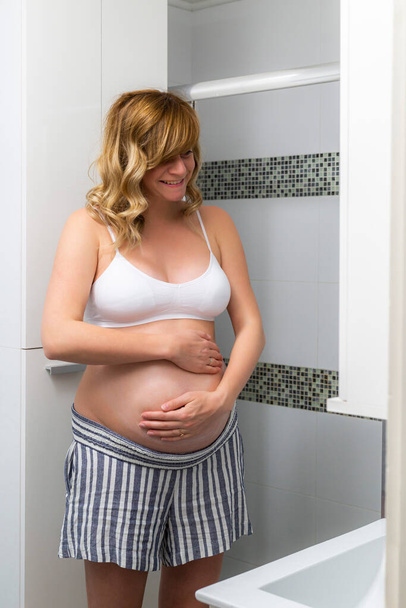 浴室に立つ笑顔で幸せな妊娠中の白人女性。両手でお腹にクリームをこすりつけてる. - 写真・画像