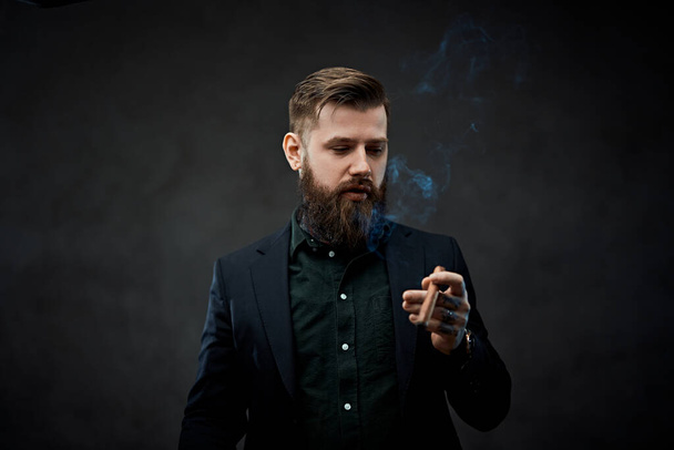 Жестокий молодой бизнесмен курит сигару в темной студии
. - Фото, изображение