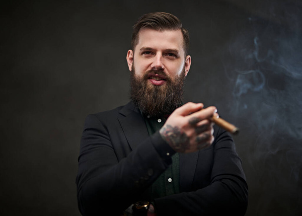 Brutal young businessman smokes a cigar in a dark studio. - Photo, Image