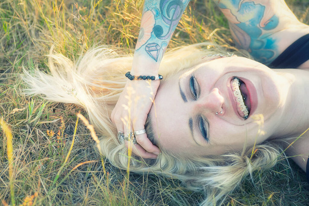 Blonde woman with tattoos and braces laughing in the grass - Фото, зображення