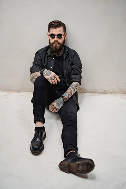 Elegante barbudo con tatuajes en las manos y el cuello sentado en el suelo
 - Foto, Imagen