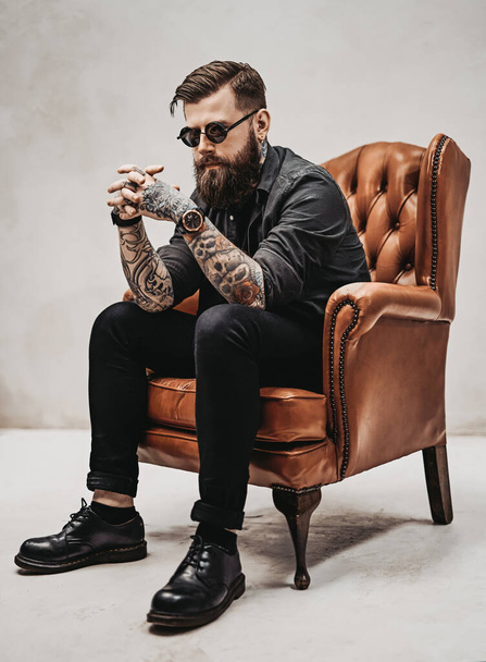 Крутой бородатый молодой человек сидит на винтажном стуле в студии
 - Фото, изображение