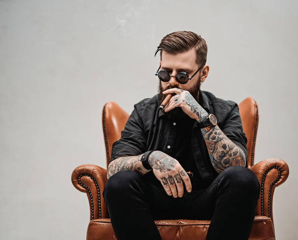Tattooed bearded man with stylish haircut in sunglasses who smokes a cigar while sitting on a vintage chair in studio - 写真・画像