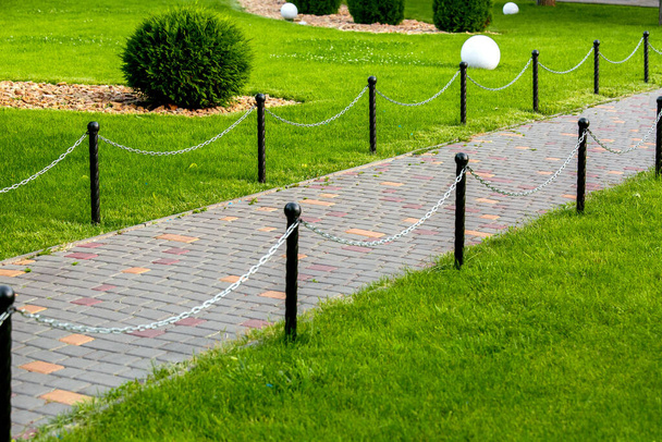 stone walkway and tiles in the park with a fence made of black columns and a chain stretched between the supports in the garden with green grass of the backyard close up nobody. - Photo, image