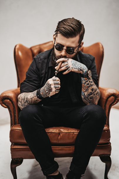 Homem de barba tatuado com corte de cabelo elegante em óculos de sol que fuma um charuto enquanto se senta em uma cadeira vintage em estúdio
 - Foto, Imagem