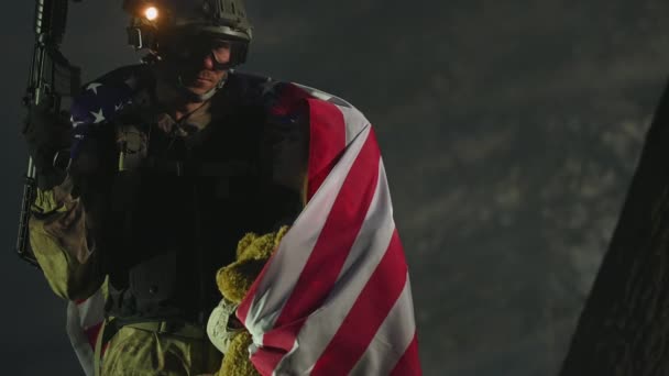 Giovane ragazzo con bandiera americana avvolta in giro tenendo orsacchiotto ed essendo abbracciato dal padre soldato in uniforme e arma dell'esercito pieno nella mano destra. - Filmati, video