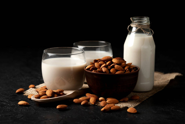 almond milk in glasses and a bottle with nuts on a dark background, a healthy dairy product without lactose on a black table, a vegetarian drink - Foto, afbeelding