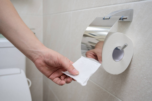Une main tend la main pour du papier toilette dans une salle de bain carrelée beige. - Photo, image
