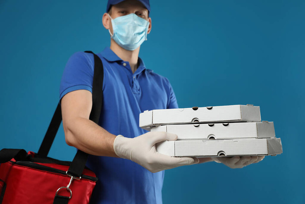 Koruyucu maskeli bir kurye ve mavi arka planda pizza kutuları tutan eldivenler. Coronavirus karantinası sırasında yiyecek teslimatı - Fotoğraf, Görsel