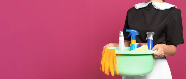 Young chambermaid holding plastic basin with detergents on color background, closeup view with space for text. Banner design - Foto, Bild