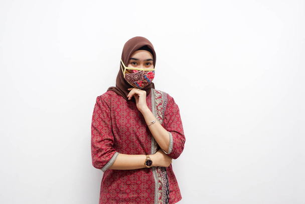 Nuori kaunis aasialainen nainen, joka pukeutuu hijabiin ja batiikkiin, hymyilee ja poseeraa valkoisella taustalla. Hän käyttää myös batiikkinaamiota suojellakseen häntä 19-vuotiaalta ja katsoo kameraan. - Valokuva, kuva