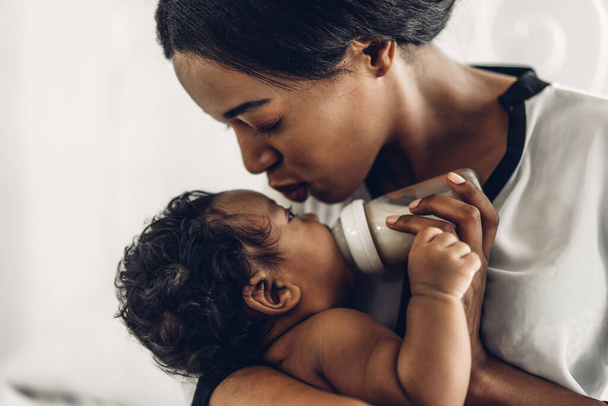 Portrait of enjoy happy love family african american mother playing with adorable little african american baby.Mom feeding bottle of milk to baby cute son in a white  bedroom.Love of black family concept - Photo, Image