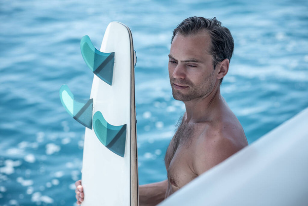 Portrait of handsome man with blue eyes, surfer holding surf board with blue fins and blue ocean on background - Foto, Imagen