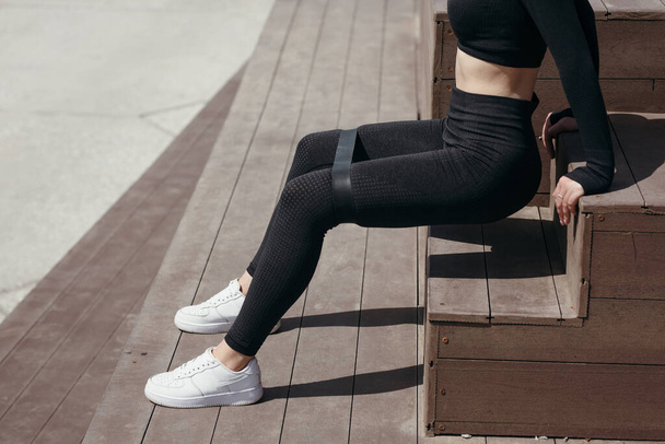 Closeup of girl sitting on a bench and doing exercises for legs with an expander to strengthen the muscles of the thighs and legs. - 写真・画像