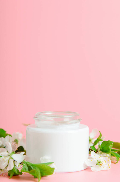 Creme de cara em jarro branco em um contexto rosa com flores brancas. Conceito cosméticos naturais, beleza orgânica. Espaço de cópia
 - Foto, Imagem