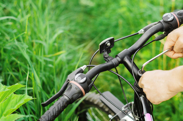 Человек откручивает болты шестигранным гаечным ключом на руле горного велосипеда на фоне травы. Ремонт на лесной дороге. Крупный план
. - Фото, изображение