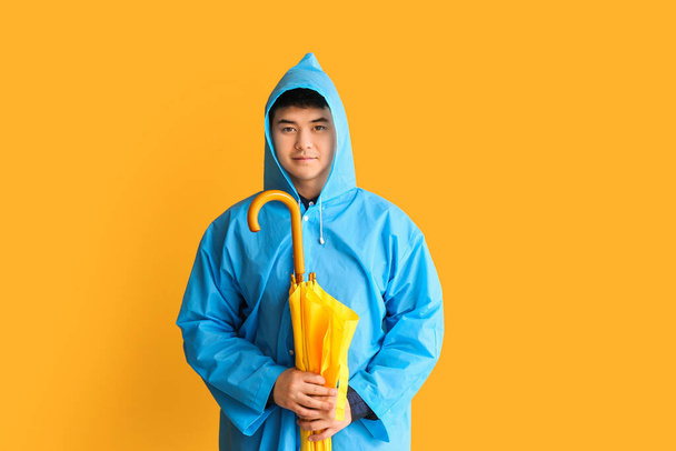 Young Asian man in raincoat and with umbrella on color background - Foto, immagini